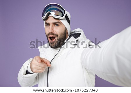 Close up of shocked skier man in windbreaker jacket ski goggles mask doing selfie shot on mobile phone point on camera spend weekend in mountains isolated on purple background. People hobby concept