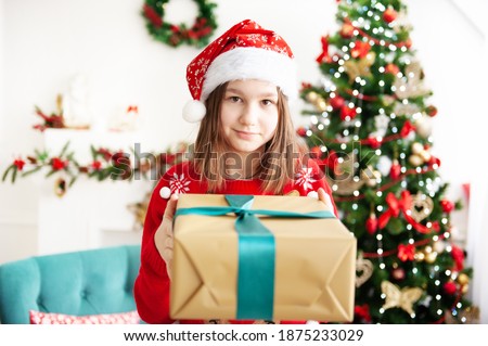 A young teenage girl in a Christmas cap and a red sweater with a large gift is sitting in a cozy chair next to the Christmas tree. Congratulation 