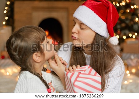 Young winsome mother and daughter with gift box looking at each other, wants to kiss her cute little girl, Christmas Eve at home, posing near fireplace.