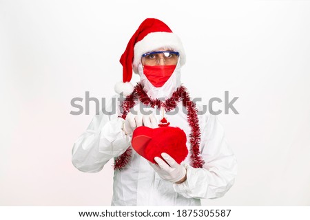 Health worker wearing a Santa hat. He is wearing an antivirus overall. He wears a red medical mask on his face. He is holding a gift box in the shape of a heart. Isolated background.