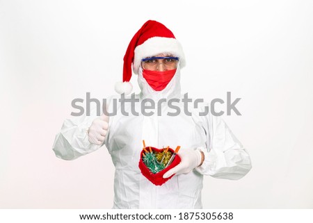 Health worker wearing a Santa hat. He is wearing an antivirus overall. He wears a red medical mask on his face. He is holding a gift box in the shape of a heart. Isolated background.