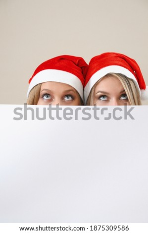 Two funny girls in Santa's red hat with big white empty card looking up. Happy holiday concept. New Year party