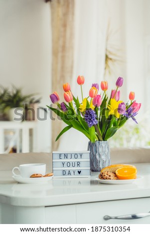 Good morning concept. Romantic breakfast - fresh flowers, cup of hot drink, cookies, orange, lightbox with message Enjoy your day on marble table with light interior view. Vertical card. Copy space.