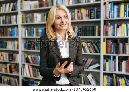 Portrait of caucasian amazing mature business woman or teacher in a formal stylish suit, holding and using mobile phone and looking away with smile