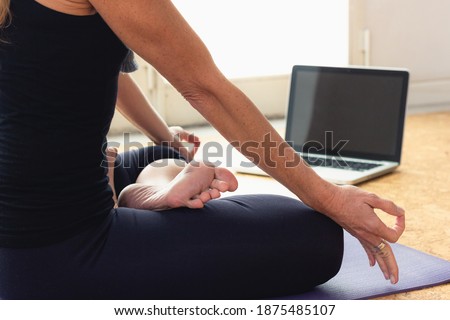 Body of slim woman sitting in padmasana in front of laptop at home. Female yogi on black outfit doing lotus pose indoors. Online yoga class concept. Ring language translation: Honoring Great Ganesha