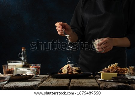 Close-up view of chef pours coconut for serving waffles on rustic wooden table with ingredients on dark blue background. Backstage of cooking sweet dessert for breakfast. Frozen motion.