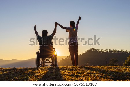 Disabled handicapped young man in wheelchair raised hands with his care helper in sunset.Silhouette Royalty-Free Stock Photo #1875519109