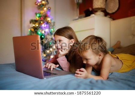 Girls children in bedroom are looking at screen near Christmas tree in real interior, concept of Christmas sales and interesting cartoons. siblings on the bed with blanket lying looking laptop