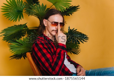 An attractive man with long hair and a mustache in a red plaid 80s disco shirt sits on a chair against a yellow background. Guy in fashionable sunglasses on the background of a green palm tree