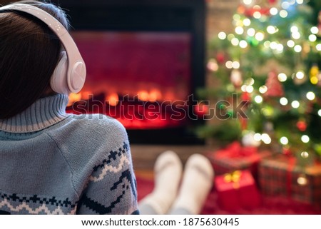 Alone sad woman in headphones sitting and warming at winter evening near fireplace flame and  christmas tree.