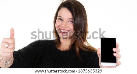 young woman thumbs up holding cell phone with black empty copyspace screen posing in white blank studio wall