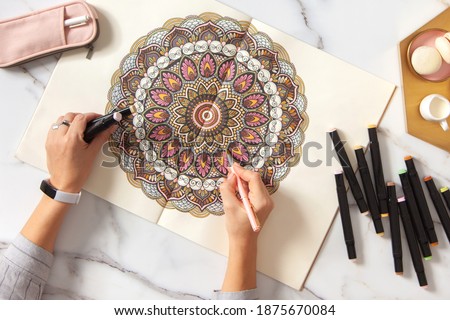 Woman drawing and coloring mandala in sketchbook with colorful markers while having breakfast with coffee and macarons on white marble table. Stress relieving trend by painting. Art therapy. Leisure. Royalty-Free Stock Photo #1875670084