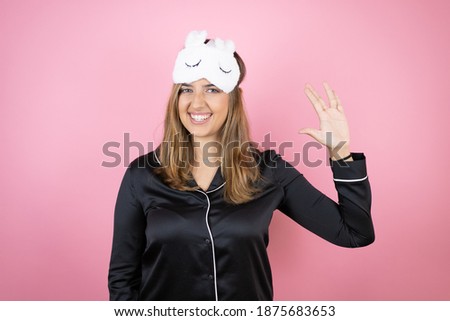 Young caucasian woman wearing sleep mask and pajamas over isolated pink background doing hand symbol