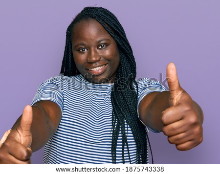 Young black woman with braids wearing casual clothes approving doing positive gesture with hand, thumbs up smiling and happy for success. winner gesture. 