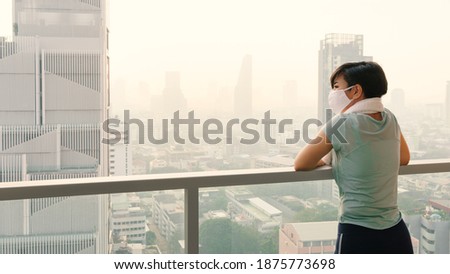 Beautiful Asian woman wearing medical face mask look at downtown skyscrapers in Bangkok city covered with thick smog. Sign of poor air quality, high risk of PM 2.5 pollution with serious health effect Royalty-Free Stock Photo #1875773698