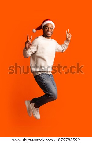 Christmas Party. Playful Black Man In Santa Hat Jumping And Gesturing Peace Over Orange Background In Studio, Having Fun And Showing V-Sign With Both Hands, Celebrating Xmas Holidays, Free Space