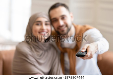 Cheerful Muslim Couple Watching Movie On TV Sitting Together On Sofa At Home, Focus On Television Remote Control. Arab Husband And Wife Switching Channels Looking At Camera. Shallow Depth