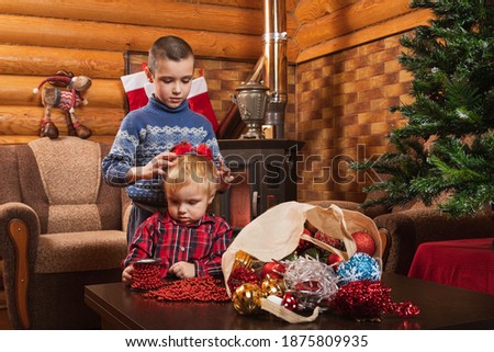 A little boy in a warm knitted sweater puts deer horns on his brother in a country house, in the background there is a decorated Christmas tree, a lighted fireplace, socks for gifts are hung. 