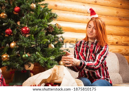 A cheerful woman in a plaid shirt hangs a beautiful shiny ball on a Christmas tree oon the background of a lighted fireplace, hanging socks for gifts. Christmas and new year home preparation