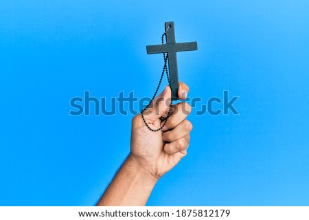 Hand of hispanic man holding christian cross over isolated blue background.