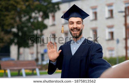 education, graduation and people concept - happy smiling male graduate student in mortar board and bachelor gown taking selfie and waving hand over school background