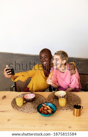 Beautiful young lesbian couple is doing selfie using a smart phone and smiling while having a breakfast at home