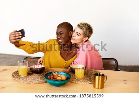 Beautiful young lesbian couple is doing selfie using a smart phone and smiling while having a breakfast at home