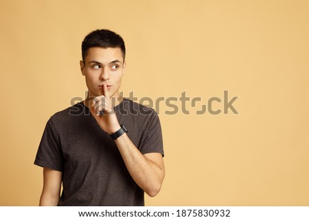 Secret, offer, interesting advertisement and gossip. Mysterious young male student with smart watch shows hand sign silence and looks towards empty space isolated on light sand background, studio shot