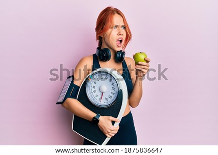 Young beautiful redhead woman holding weight machine to balance weight loss angry and mad screaming frustrated and furious, shouting with anger. rage and aggressive concept.  Royalty-Free Stock Photo #1875836647