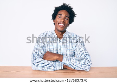 Handsome african american man with afro hair wearing casual clothes sitting on the table happy face smiling with crossed arms looking at the camera. positive person. 