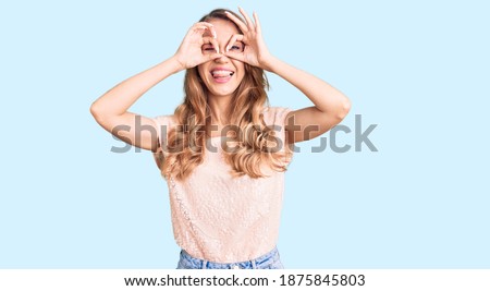 Young beautiful caucasian woman with blond hair wearing casual clothes doing ok gesture like binoculars sticking tongue out, eyes looking through fingers. crazy expression. 