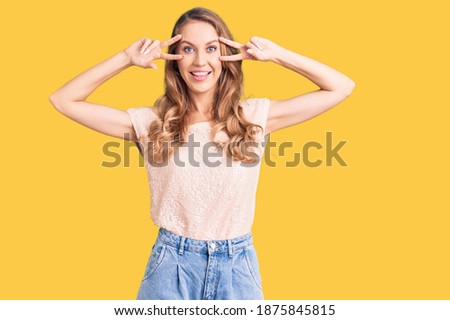 Young beautiful caucasian woman with blond hair wearing casual clothes doing peace symbol with fingers over face, smiling cheerful showing victory 