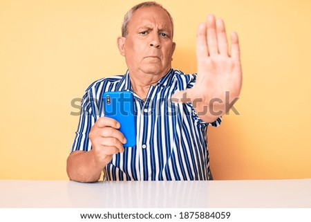 Senior handsome man with gray hair using smartphone sitting on the table with open hand doing stop sign with serious and confident expression, defense gesture 