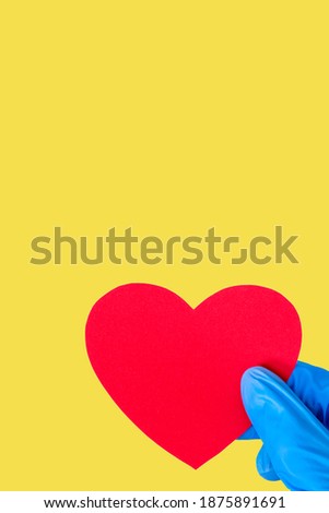 Valentine's day 2021 concept. Hand in medical blue gloves holds a red heart shape on a yellow Illuminating background, copy space, vertical frame, mock up. Love, health concept