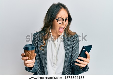 Young brunette woman using smartphone and drinking a cup of coffee angry and mad screaming frustrated and furious, shouting with anger. rage and aggressive concept. 