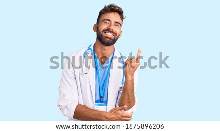 Young hispanic man wearing doctor uniform and stethoscope with a big smile on face, pointing with hand and finger to the side looking at the camera. 
