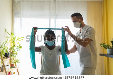 Young african woman wearing a mask doing rehabilitation exercises of the arms with a rubber band with a physiotherapist wearing a mask and a uniform in a clinic Royalty-Free Stock Photo #1875915397