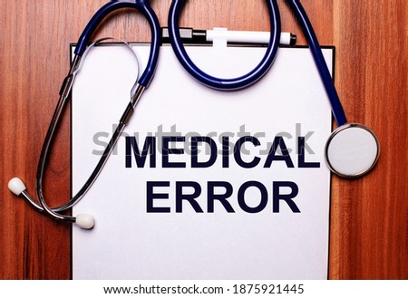 The word MEDICAL ERROR is written on white paper on a wooden background near a stethoscope and black-framed glasses. Medical concept