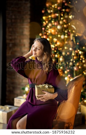 The girl sits in a chair by the Christmas tree. The girl is holding a gift in her hands. Image with selective focus.