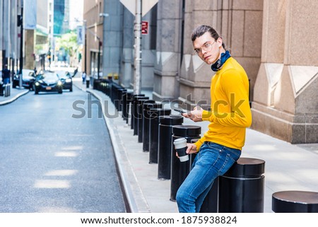 Young Hispanic American with hair bun, wearing glasses, yellow long sleeve T shirt, blue jeans, small black scarf around neck, holding cup of coffee, sitting on street in New York City, texting.
