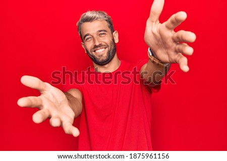 Young handsome blond man wearing casual red t-shirt standing over isolated red background looking at the camera smiling with open arms for hug. Cheerful expression embracing happiness.
