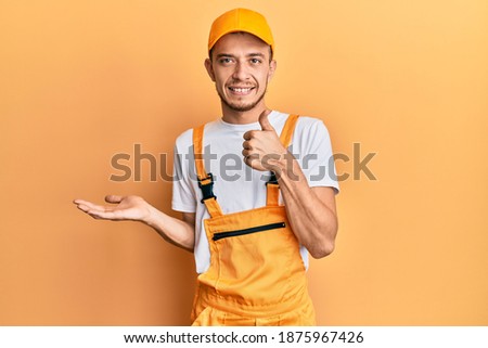 Hispanic young man wearing handyman uniform showing palm hand and doing ok gesture with thumbs up, smiling happy and cheerful 