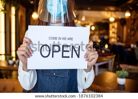 Small business owner smiling while turning the sign for the reopening of the place after the quarantine due to covid-19. Happy woman standing at her restaurant or coffee shop gate with open signboard.