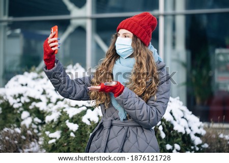 Female blogger in face mask is making video talking for her social page at the street in winter time. Woman look around on the winter street city. Being online, social networks.