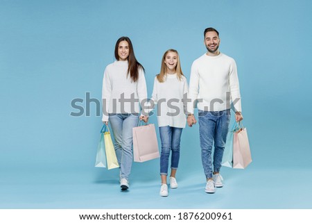 Full length of funny young parents mom dad with child kid daughter teen girl in sweaters hold package bags with purchases after shopping isolated on blue background. Family day parenthood concept Royalty-Free Stock Photo #1876200961