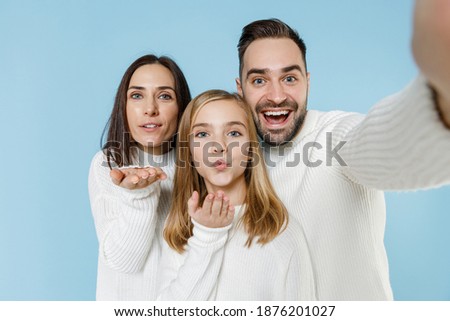 Close up of pretty young parents mom dad child kid daughter teen girl in sweaters doing selfie shot on mobile phone blowing sending air kiss isolated on blue background. Family day parenthood concept