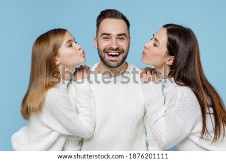 Cheerful young happy parents mom dad with child kid daughter teen girl in casual white sweaters kissing on cheeks isolated on blue background studio portrait. Family day parenthood childhood concept
