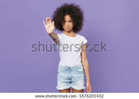 Little african american kid girl 12-13 years old in white t-shirt isolated on violet wall background studio portrait. Childhood lifestyle concept. Mock up copy space. Showing stop gesture with palm Royalty-Free Stock Photo #1876201432