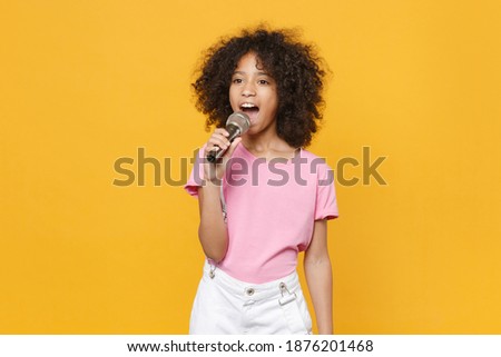 Charming little african american kid girl 12-13 years old in pink t-shirt isolated on yellow wall background studio portrait. Childhood lifestyle concept. Mock up copy space. Sing song in microphone Royalty-Free Stock Photo #1876201468