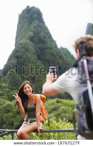 Couple taking photos having fun lifestyle hiking on Hawaii in outdoor activity. Woman and man hiker taking photo pictures with smart phone camera. Iao Valley State Park, Wailuku, Maui, USA.